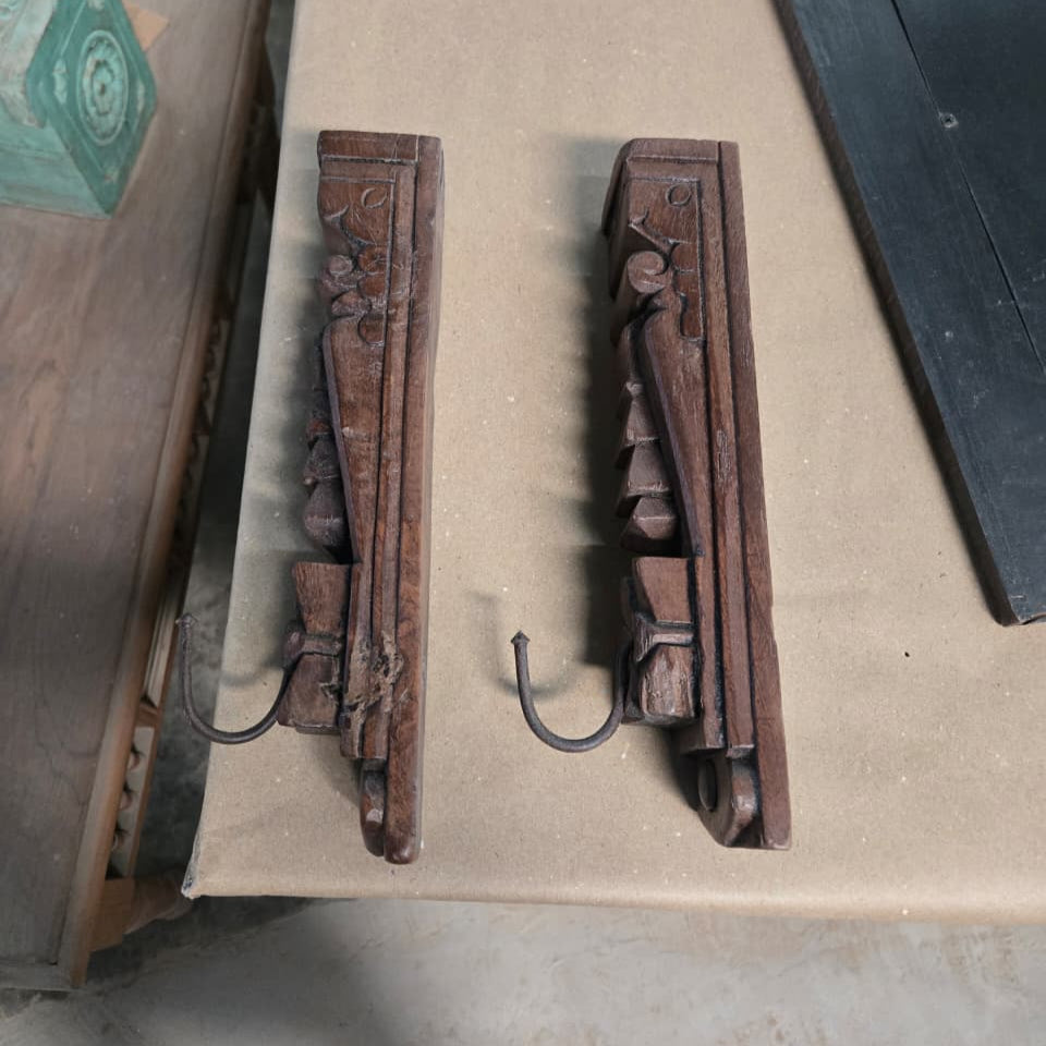 Introducing the Ankush Coat Hooks: a set of two carved wooden wall hooks featuring ornate geometric and floral details, showcased on a beige surface. Each hook is crafted with exquisite attention to detail and includes a sturdy metal hanger extending from the base, making them an elegant handcrafted decor accent for any space.