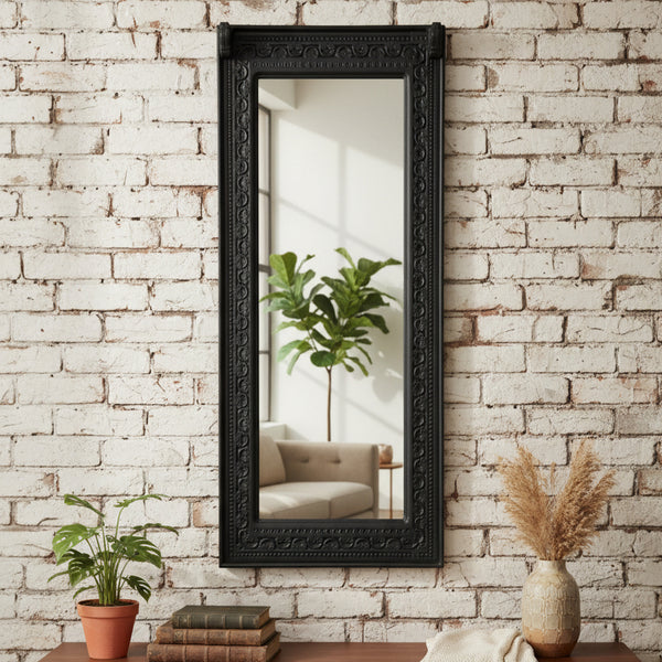 Ashva Wooden Mirror Frame with intricate carved wooden border, hanging on a white brick wall, reflecting a cozy living room with a beige sofa and green plant for stylish home decor.