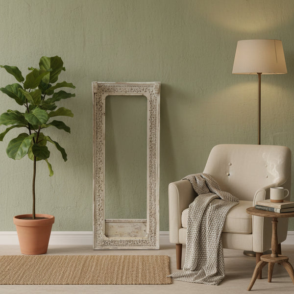 Ashva Wooden Mirror Frame with intricate carved wooden details, leaning against a sage green wall in a cozy living space featuring a cream armchair, floor lamp, and potted plant.