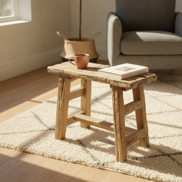 Rustic wooden stool with a handcrafted tribal design placed on a textured rug, featuring a ceramic mug and a book, next to a cozy armchair in a sunlit room.
