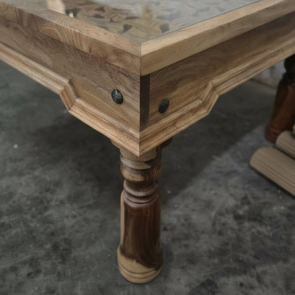 Handcrafted wooden table with carved details, glass top, and turned leg on concrete floor