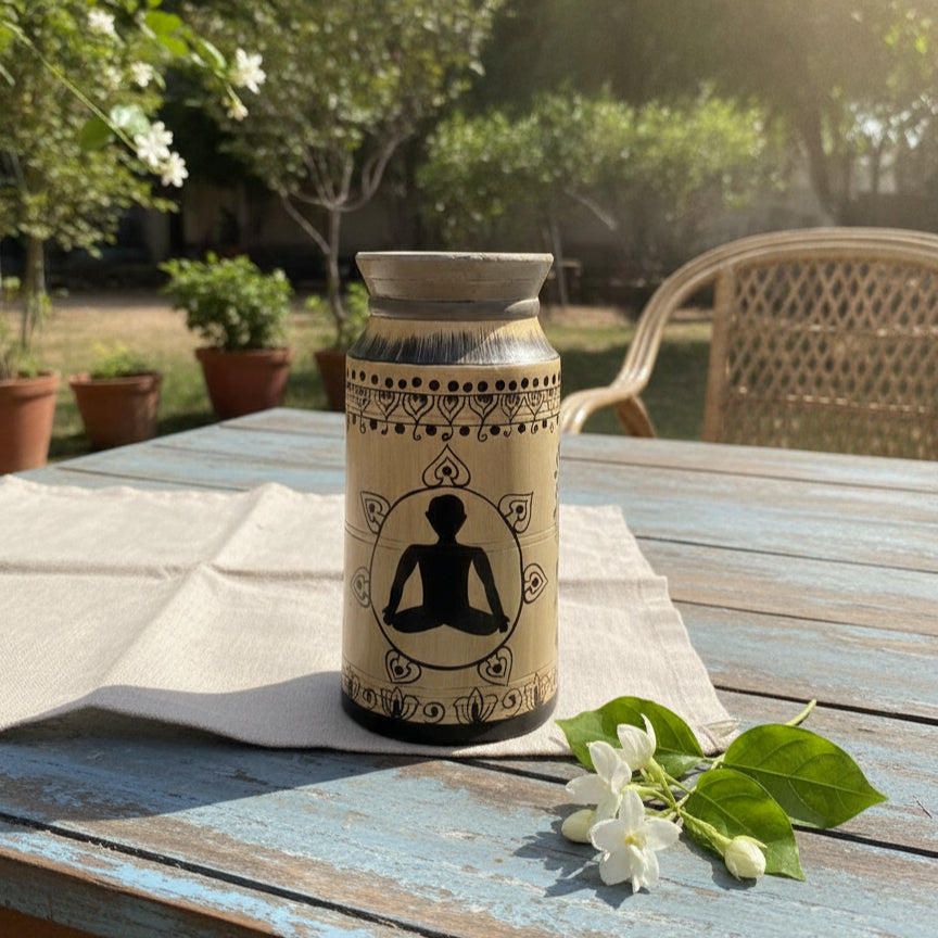Hand-painted wooden pot with a serene meditating figure and intricate tribal-inspired patterns, placed on a rustic table with fresh jasmine flowers nearby.
