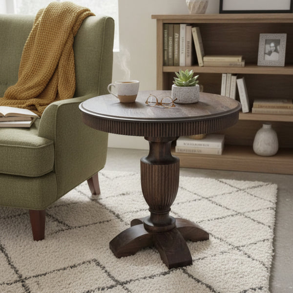 Round wooden pedestal table with carved detailing placed on a textured rug beside a green armchair with a mustard throw and a bookshelf in the background