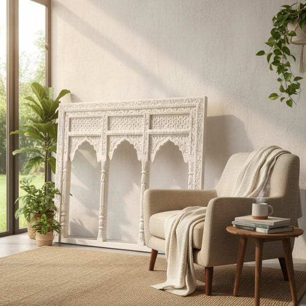 A cozy living room with a beige armchair, a small wooden table with a cup, potted plants, and the large ornate white Jharokha Wall Hanging leaning against a sunlit wall. A textured rug covers the floor.