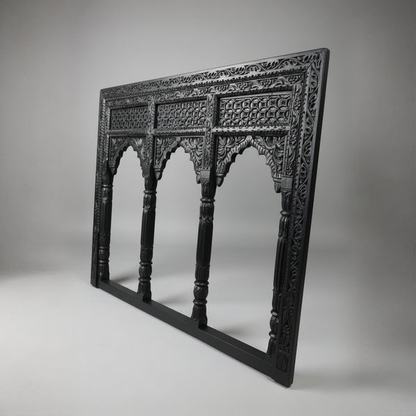 The Jharokha Wall Hanging is a carved, rectangular black wooden frame featuring intricate patterns and three arched openings that echo traditional architectural window designs. Displayed against a plain grey background, this exquisite piece of wooden décor beautifully showcases expert handicraft.