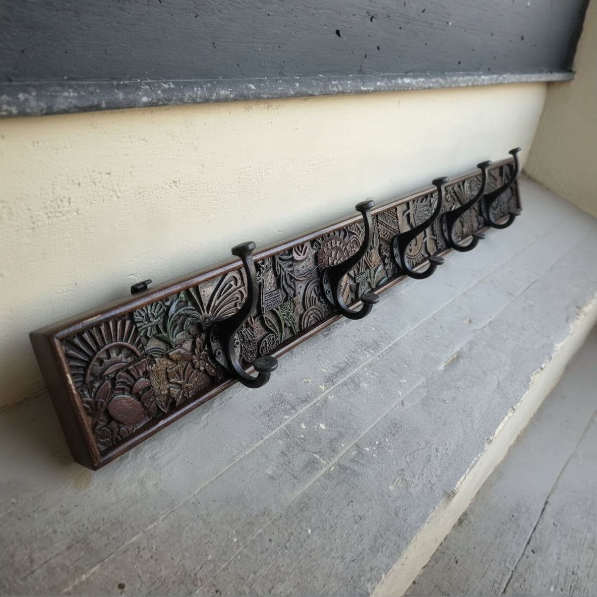 Wooden wall-mounted coat rack featuring intricate tribal hand-carved patterns with multiple black metal hooks, combining traditional decor with functional design.