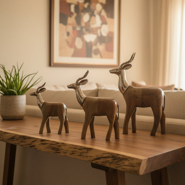 The Mriga Trio Wooden Decor, featuring three wooden antelope sculptures of different sizes, is elegantly displayed on a wooden table, highlighting exquisite craftsmanship. A potted plant sits nearby, enhancing the decor, while a blurred abstract painting adorns the wall in the background.