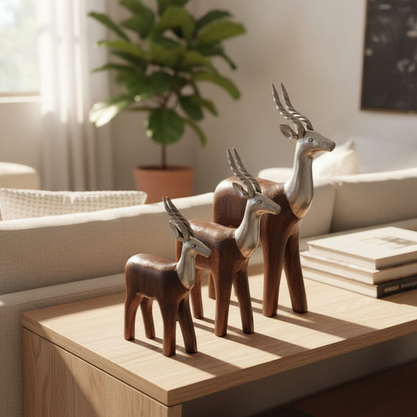 The Mriga Trio Wooden Decor, featuring three exquisitely crafted wooden antelope sculptures in varying sizes, sits atop a light wood table. A beige sofa, potted plant, and books complete the sunlit modern living room—creating an inviting space that blends artistry with comfort.