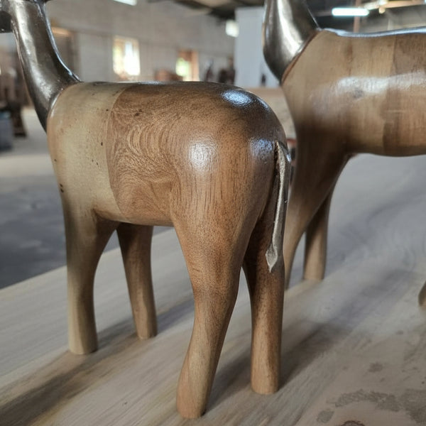 Three Mriga Trio Wooden Decor figurines, featuring smooth, polished wooden bodies and elegant thin metal tails, are displayed on a light-colored wooden surface in a sunlit room. These beautifully crafted pieces showcase the fine artistry of handcrafted decor.
