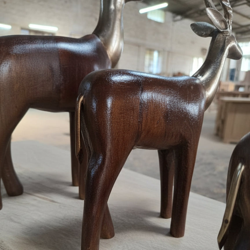 The Mriga Trio Wooden Decor features two polished wooden deer figurines with metallic heads and antlers, beautifully displayed indoors to highlight exquisite wooden craftsmanship and fine handicraft, set against a backdrop of blurred wooden furniture and a workshop setting.
