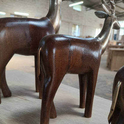 The Mriga Trio Wooden Decor features two polished wooden deer figurines with metallic heads and antlers, beautifully displayed indoors to highlight exquisite wooden craftsmanship and fine handicraft, set against a backdrop of blurred wooden furniture and a workshop setting.