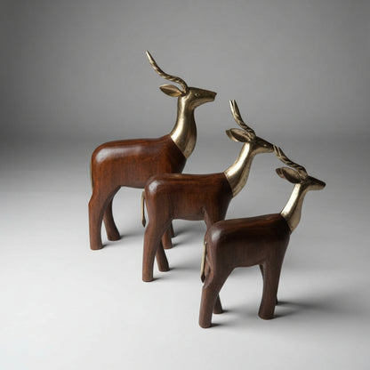 The Mriga Trio Wooden Decor features three exquisitely handcrafted wooden deer figurines with gold-colored heads and antlers, standing together on a light gray surface against a plain gray background.