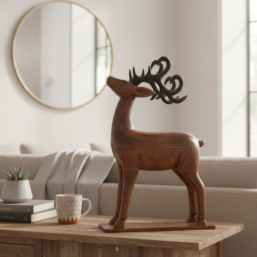 The Mriga Wooden Deer Sculpture, featuring dark antlers, stands on a table in a cozy living room beside a potted plant and a patterned mug. This charming handicraft piece adds unique decor to the space, where a round mirror hangs on the wall above a beige sofa in the background.