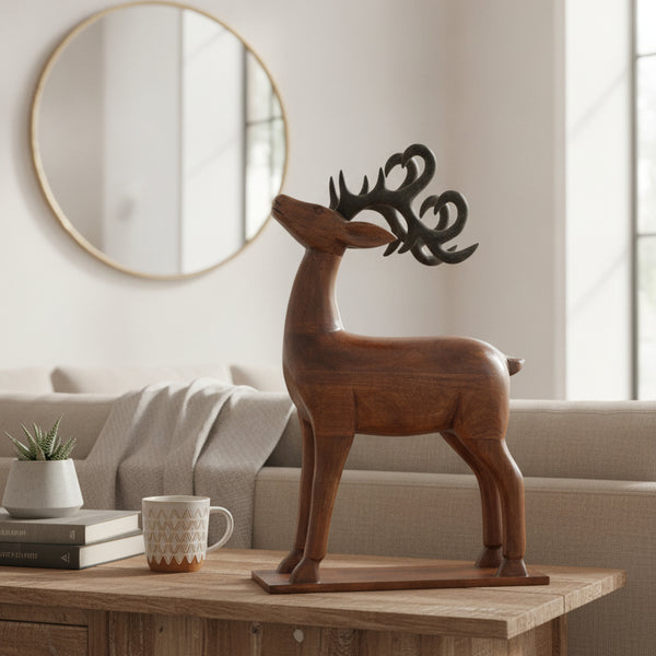 The Mriga Wooden Deer Sculpture, featuring dark antlers, stands on a table in a cozy living room beside a potted plant and a patterned mug. This charming handicraft piece adds unique decor to the space, where a round mirror hangs on the wall above a beige sofa in the background.