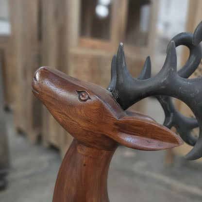 A close-up of the Mriga Wooden Deer Sculpture showcases its detailed handicraft carving, dark antlers, and smooth polished finish. Blurred wooden furniture in the background enhances its decor appeal.