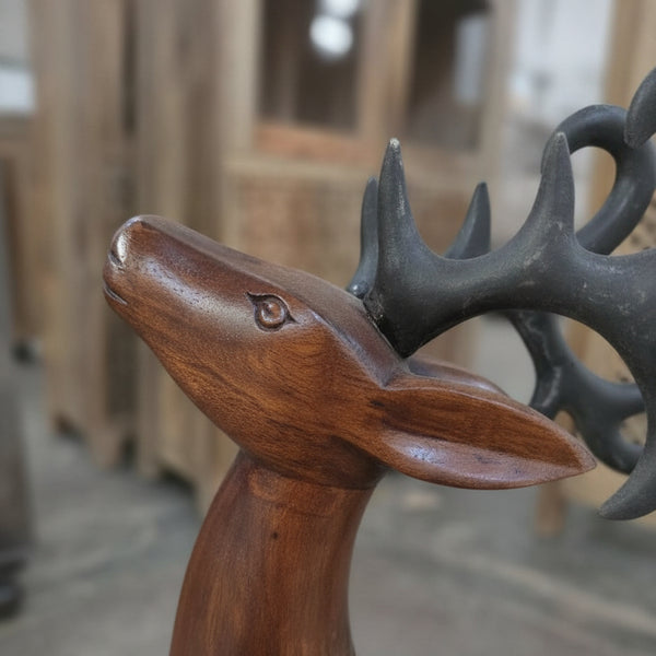A close-up of the Mriga Wooden Deer Sculpture showcases its detailed handicraft carving, dark antlers, and smooth polished finish. Blurred wooden furniture in the background enhances its decor appeal.