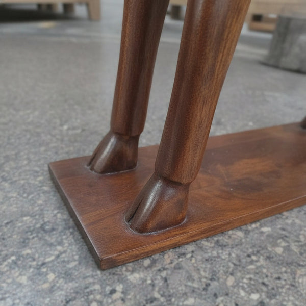 Close-up of the Mriga Wooden Deer Sculpture—a striking decor piece featuring two intricately handcrafted wooden legs, artfully shaped like deer legs and set upon a polished rectangular wooden base, displayed on a gray textured floor.