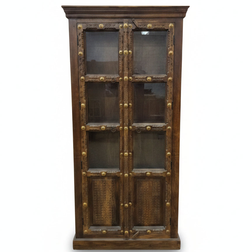 Tall wooden display cabinet with glass-paneled doors, featuring intricate tribal-style carvings and decorative brass accents, combining rustic charm and durable craftsmanship.