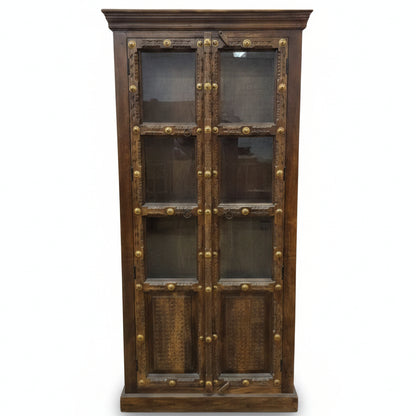 Tall wooden display cabinet with glass-paneled doors, featuring intricate tribal-style carvings and decorative brass accents, combining rustic charm and durable craftsmanship.