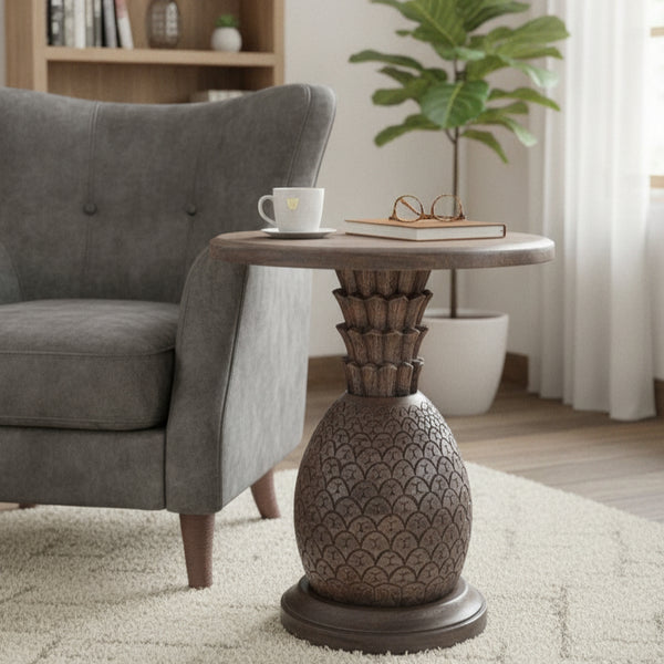 Wooden side table with intricate tribal carvings and a round top, placed next to a gray armchair in a cozy living room setting with minimal decor.