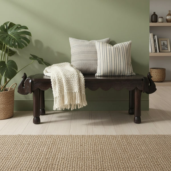 Vahana Carved Wooden Bench with dark wood finish featuring intricate tribal carvings, styled with two textured cushions and a cream knit throw, placed against a sage green wall.
